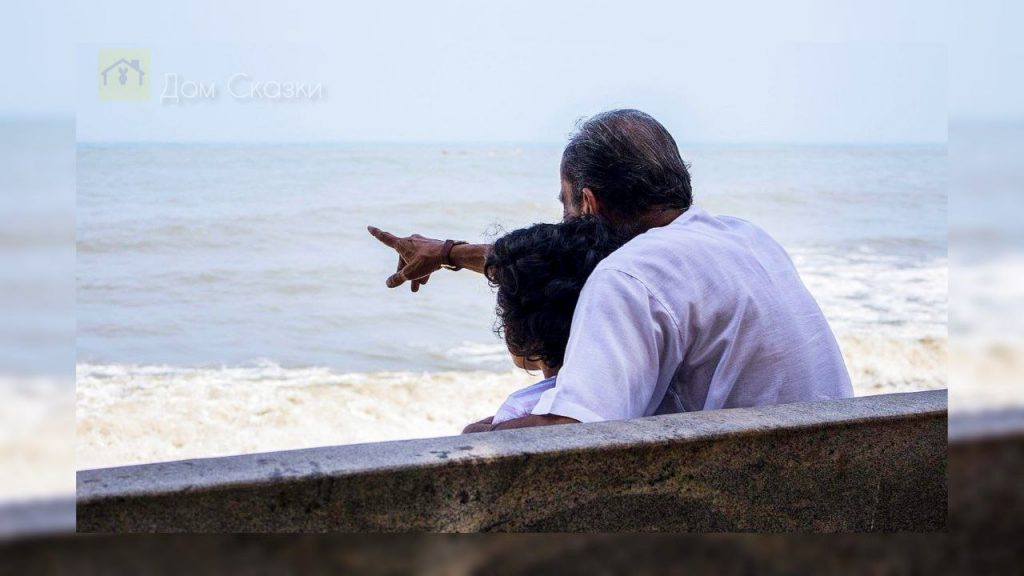 Дедушка и мальчик сидят спиной на лавочке около моря, старик показывает внуку на волны и что-то рассказывает.
