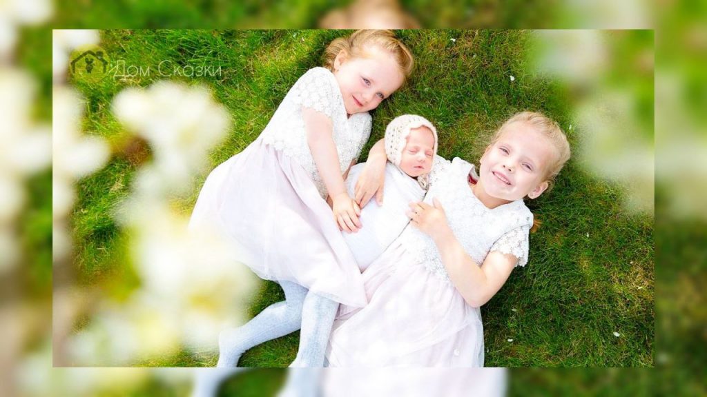 Детские вопросы часто удивительны. Две маленькие девочки в белых платьях лежат на траве, посередине их младший братик, все улыбаются и счастливы.