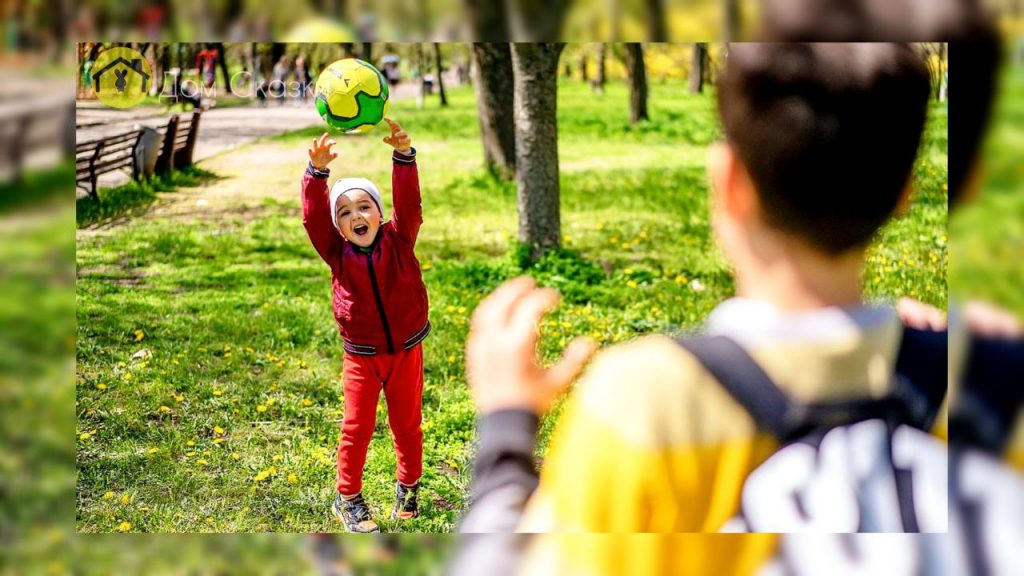 Шутливая сказка, дети играют на улице в мячик, маленькая девочка в красной толстовке и красных штанишках кидает желто-зелёный мяч мальчику, который стоит спиной.