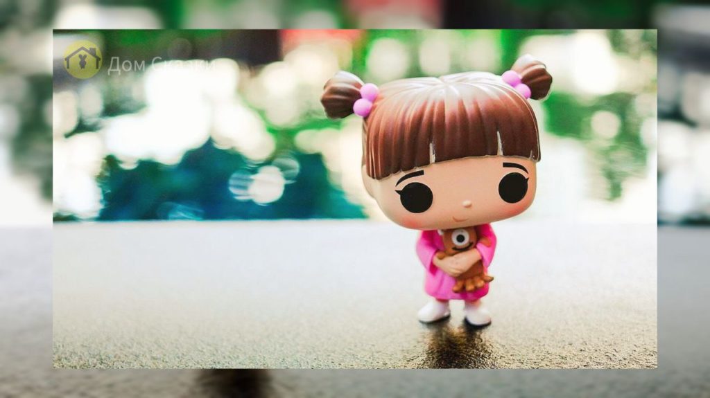 маленькая бука девочка кукла лол стоит на подоконнике. С коричневыми волосами запетёнными в хвостики, в розовом комбинезончике, с большими чёрными глазами.